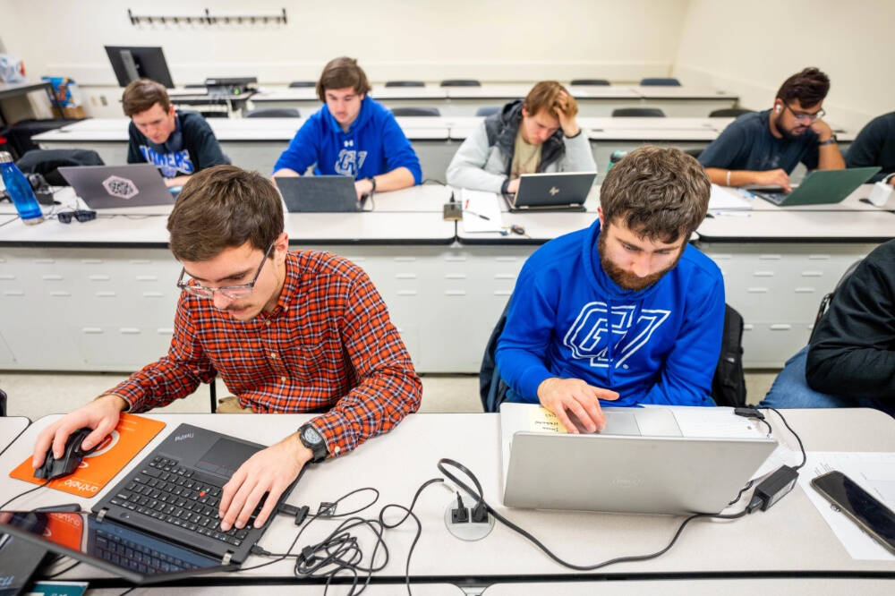 A classroom setting with students seated at long desks, each working on laptops. The students appear focused, with some typing, using a mouse, or reading. The room has a tiered layout with white walls and built-in outlets on the desks.
